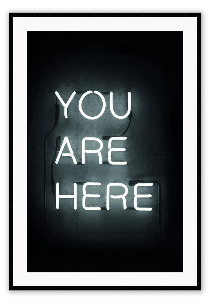 A black and white, scandi typography wall art with YOU ARE HERE neon letter lighting installation on a black wall background. 